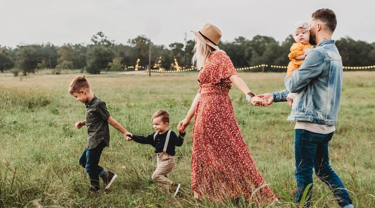 A family of five walking hand in hand through a grassy field at dusk, the father carrying a baby